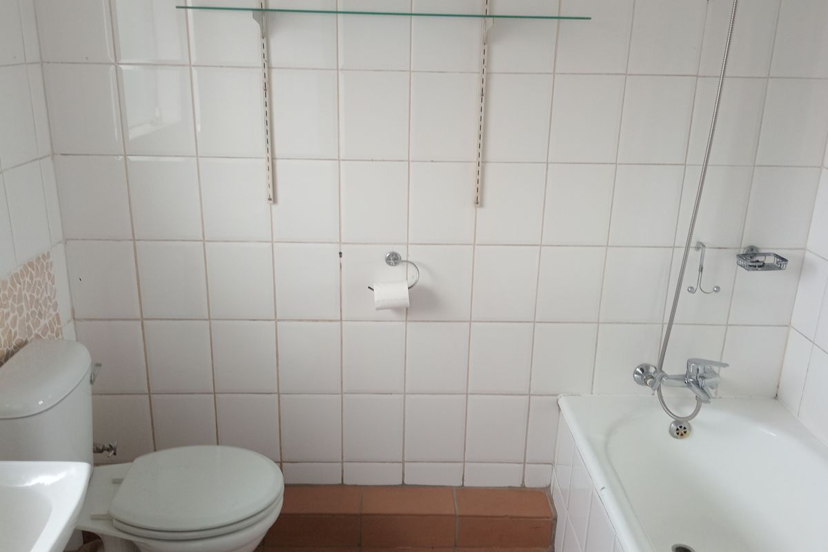 A functional bathroom featuring a standard bathtub with shower mixer, toilet, and wall-mounted shelving, finished with white wall tiles.