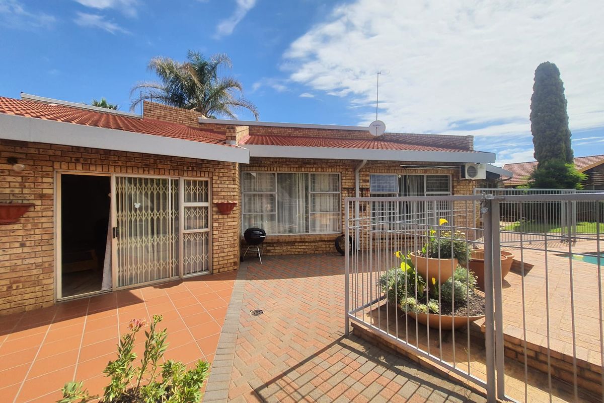 A rear exterior view of the house featuring a paved patio area, brick facade, tiled roof, and a fenced pool area.