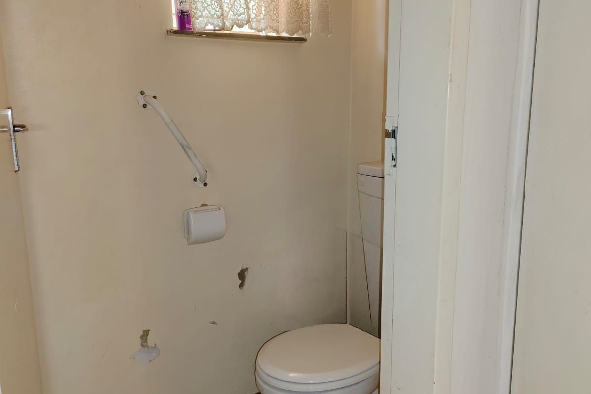 A small, dated bathroom featuring a standard toilet, tiled flooring, and a security-barred window with lace curtaining.
