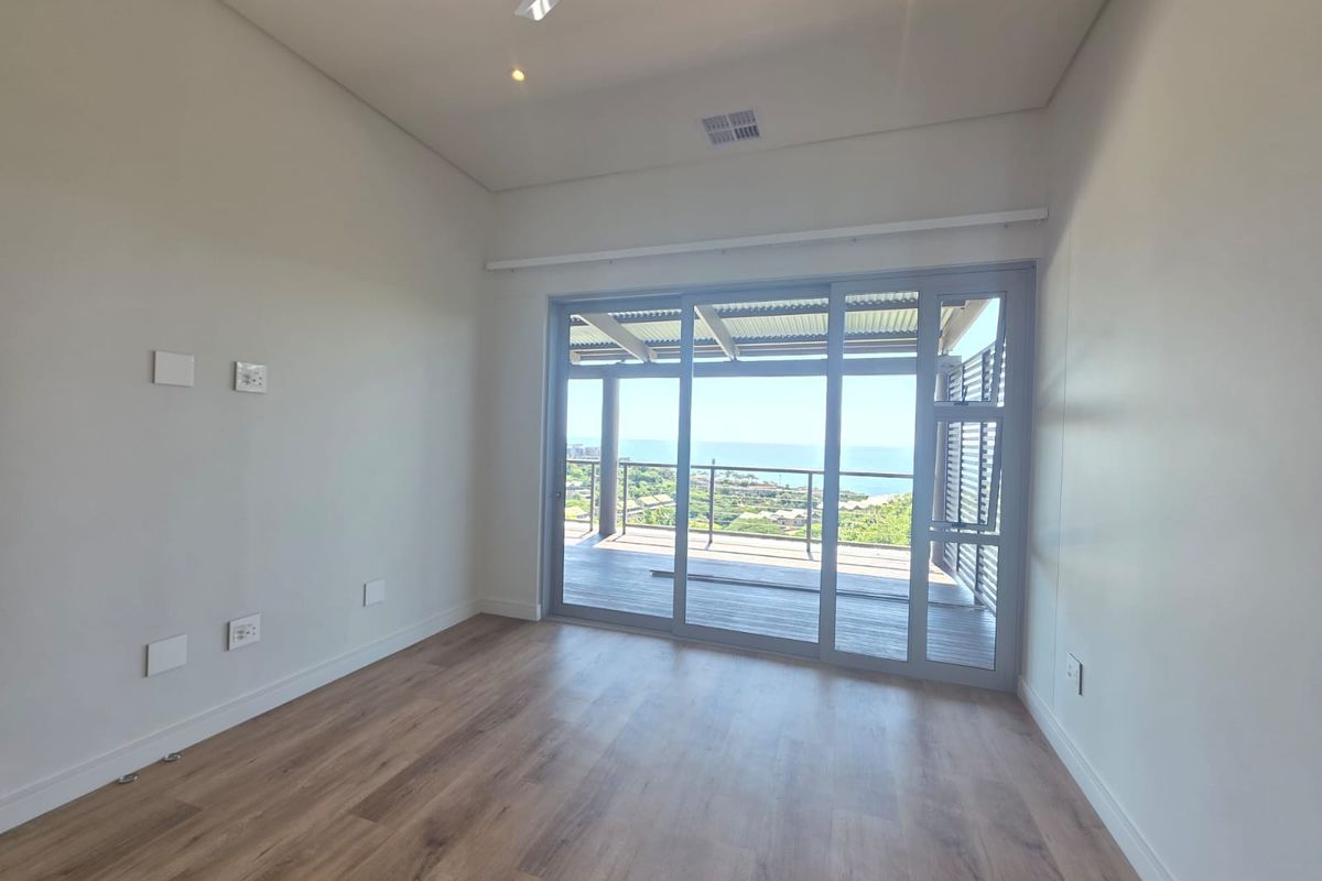 A bright, empty bedroom featuring modern wood-look flooring, a ceiling fan, and large sliding glass doors opening onto a balcony with scenic ocean views.