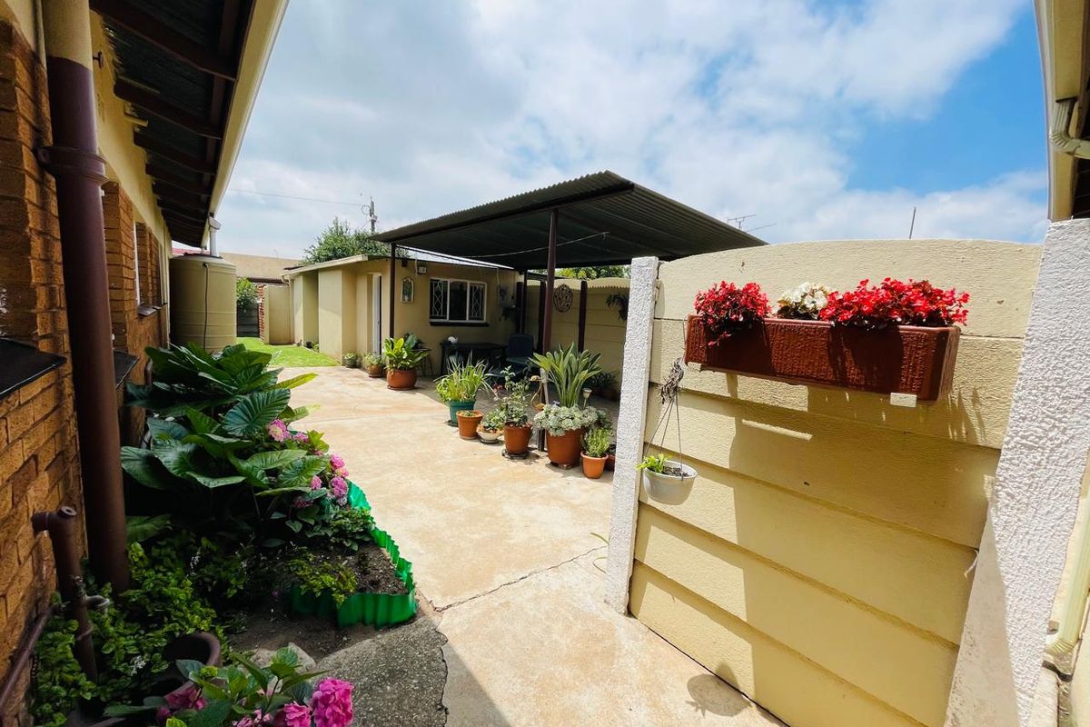 A paved rear courtyard area featuring a covered patio structure, perimeter boundary walls, and established garden beds along the side of the house.