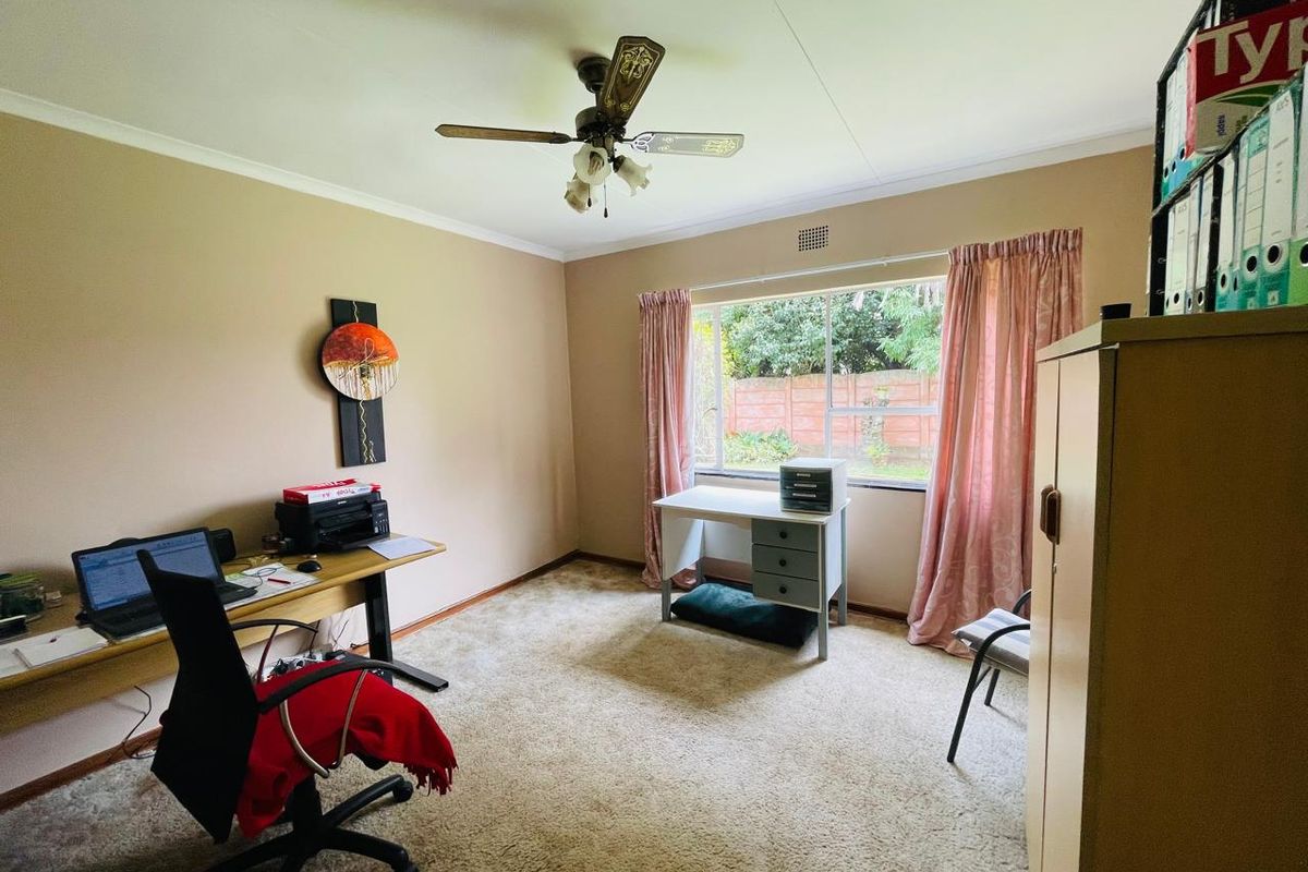 A functional bedroom or home office space featuring carpeted flooring, a ceiling fan, and a large window overlooking the rear garden area.