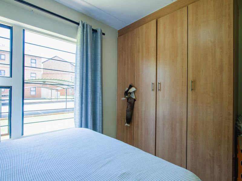A bedroom featuring built-in wooden cupboards and a large window with security bars, providing natural light and views of the apartment complex exterior.