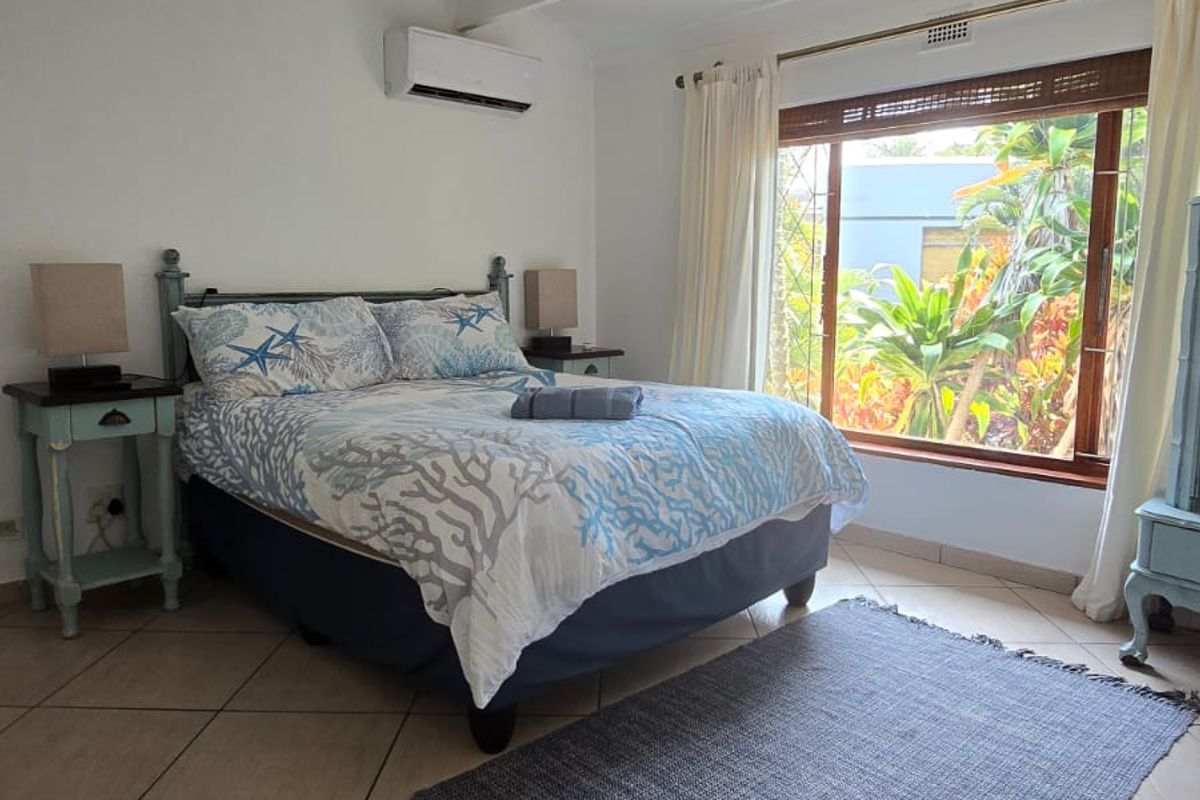 A well-lit bedroom featuring tiled flooring, a ceiling fan, and a wall-mounted air conditioning unit, with a large window overlooking the garden.