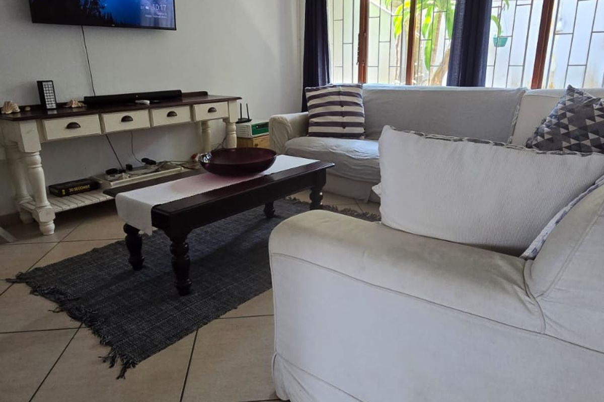 A comfortable living room featuring tiled flooring, a wall-mounted air conditioning unit, and large windows with security bars overlooking the garden.