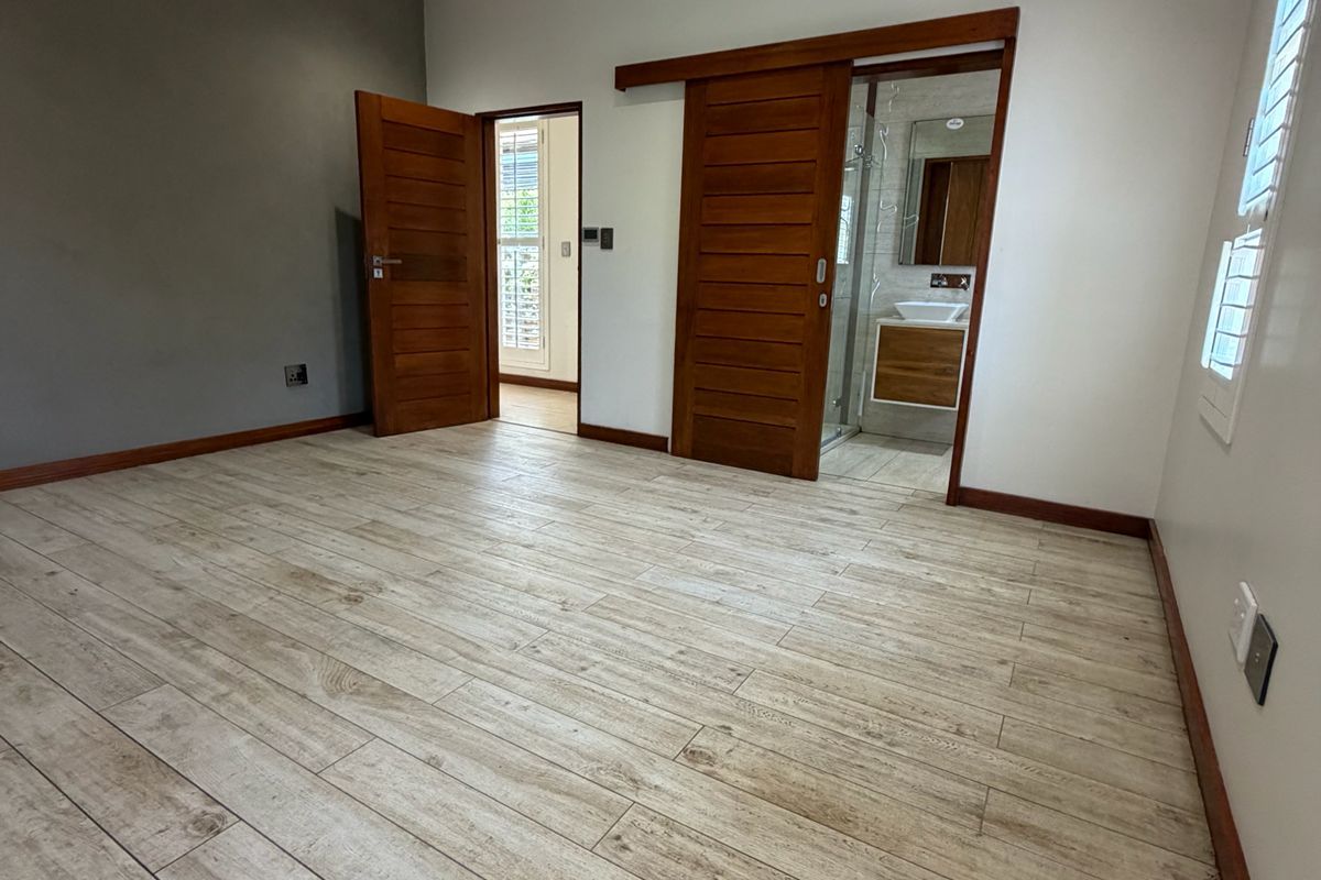 A spacious, empty bedroom featuring wood-look tile flooring, recessed lighting, and a sliding timber door leading to an en-suite bathroom.