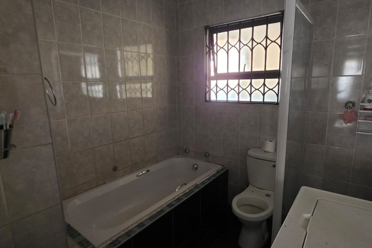 A functional bathroom featuring a built-in bathtub, a standard toilet, and tiled walls with a security-grille window.