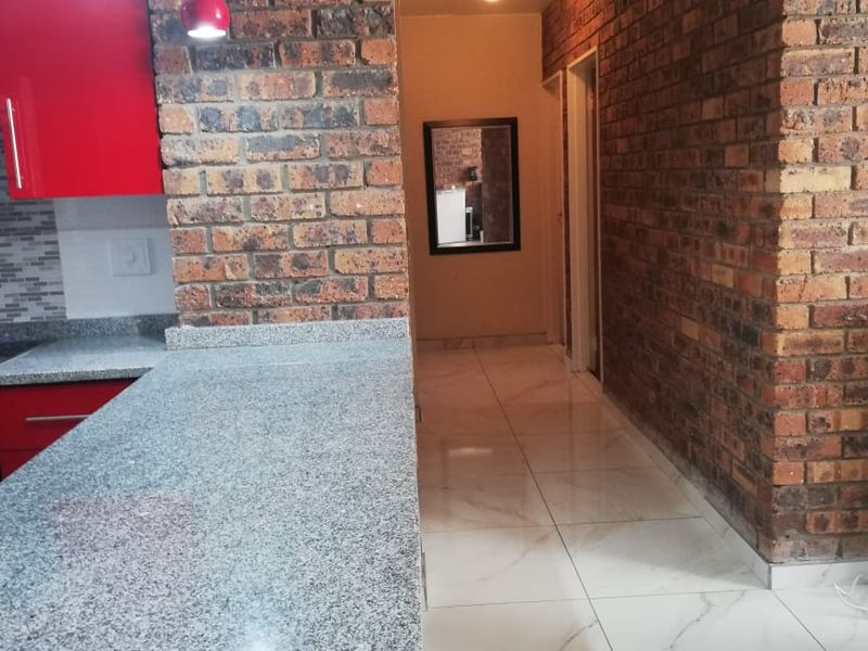 Modern kitchen featuring granite countertops, striking red cabinetry, and exposed brick walls, leading into a tiled hallway.