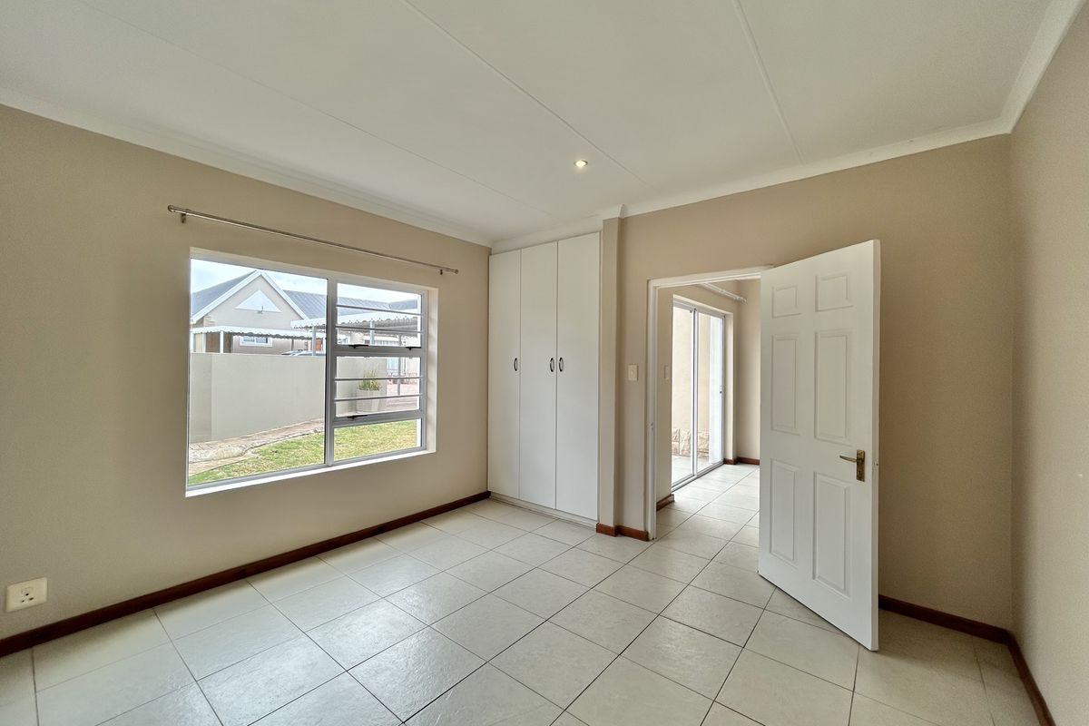 A bright, empty bedroom featuring neutral tiled flooring, built-in cupboards, and a large window overlooking the exterior garden area.