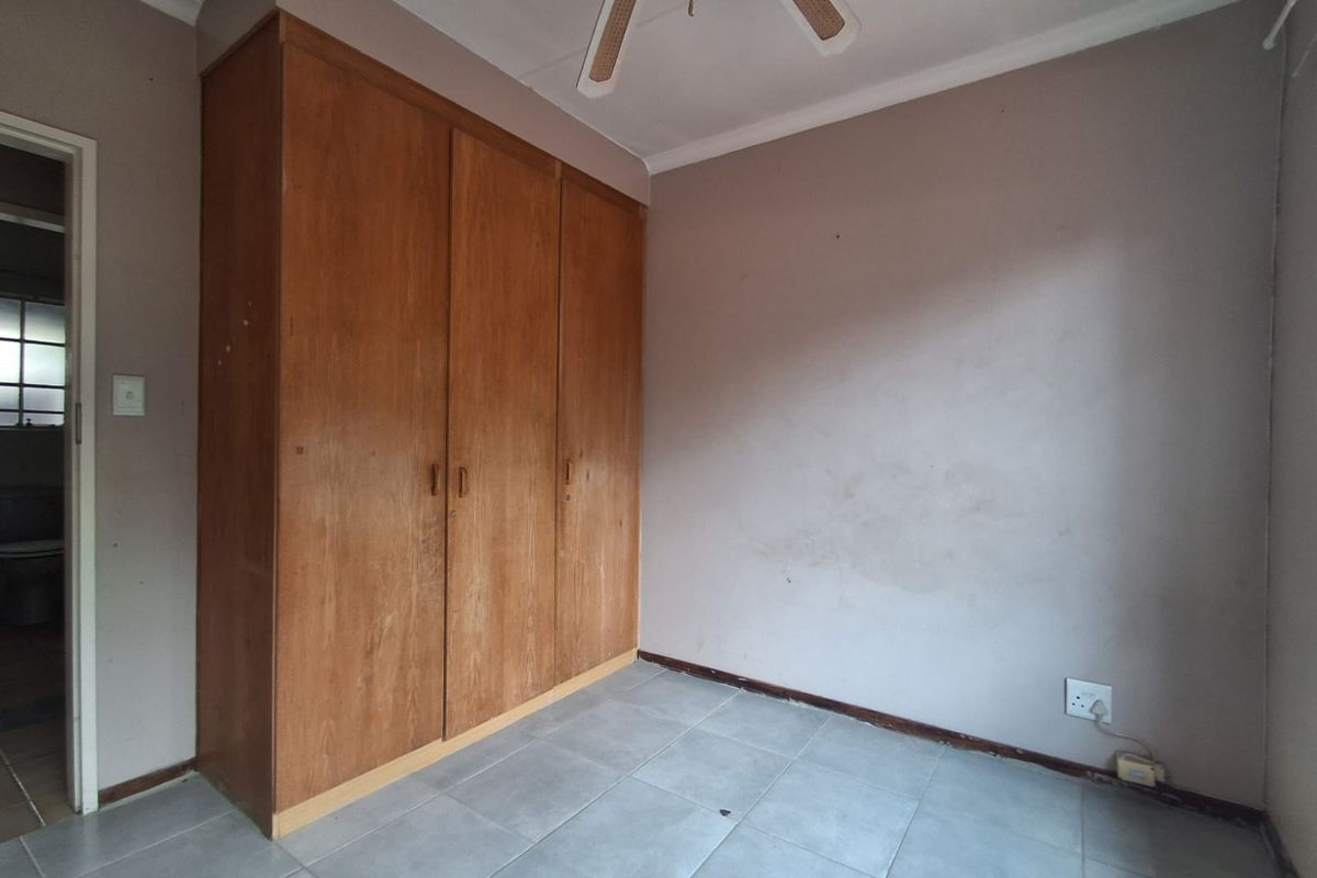 A bedroom featuring built-in wooden cupboards, tiled flooring, and a ceiling fan, presenting a dated interior in need of cosmetic updates.