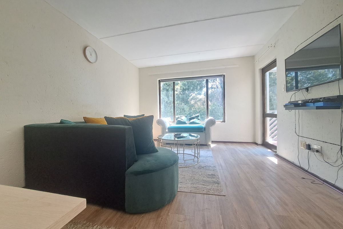 A living room featuring laminate flooring, textured walls, and a large window providing natural light, with access to an outdoor area via a glass door.