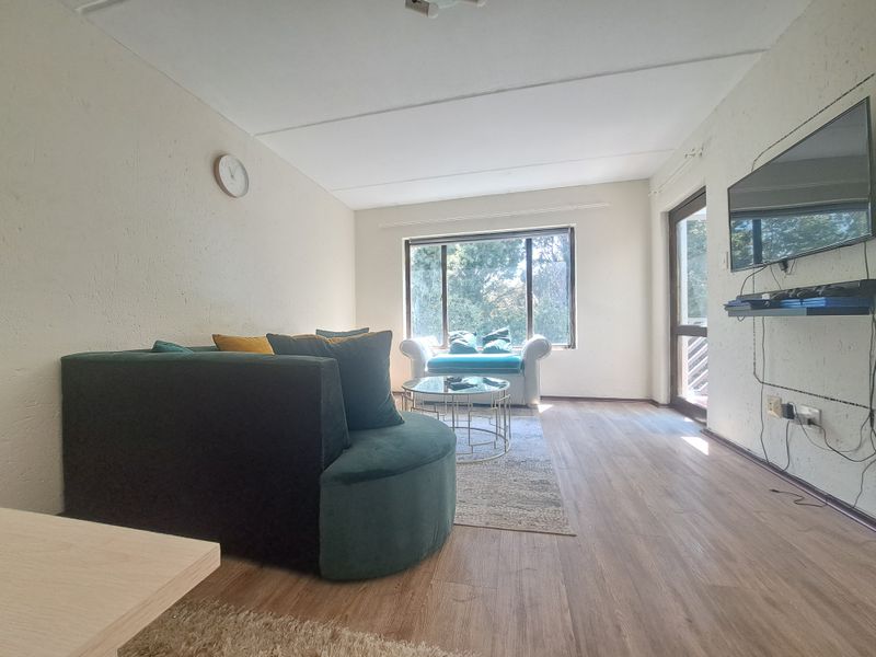 A living room featuring laminate flooring, textured walls, and a large window providing natural light, with access to an outdoor area via a glass door.