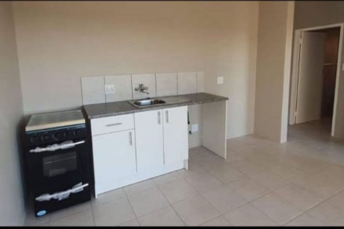 A compact, empty kitchen area featuring white cabinetry, a stainless steel sink, tiled splashback, and a freestanding stove on tiled flooring.
