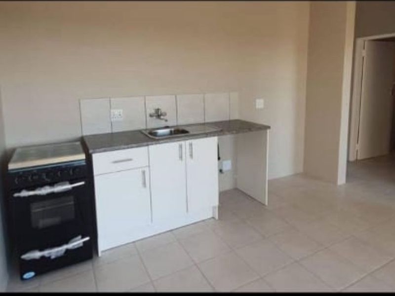 A compact, empty kitchen area featuring white cabinetry, a stainless steel sink, tiled splashback, and a freestanding stove on tiled flooring.