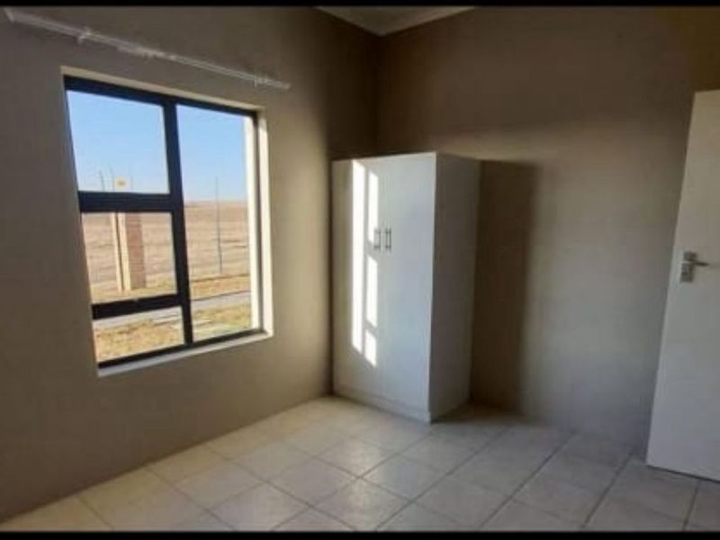A simple bedroom featuring tiled flooring, an aluminium-framed window, and a built-in cupboard, offering a functional and neutral living space.