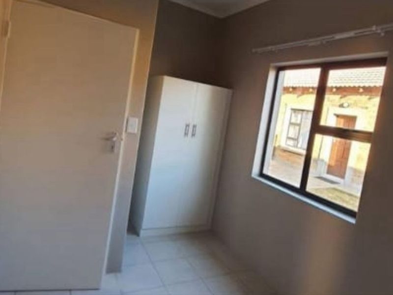A simple, neutral-toned bedroom featuring tiled flooring, a built-in wardrobe, and an aluminium-framed window overlooking the exterior.