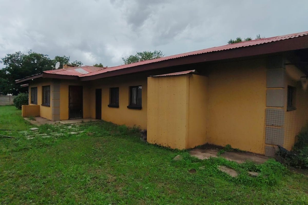 Rear view of a single-story house featuring a red corrugated metal roof, yellow exterior walls, and a large surrounding garden area.