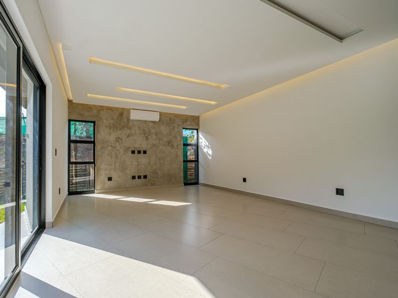A bright, modern, and empty living room featuring large format floor tiles, a textured feature wall, recessed ceiling lighting, and sliding glass doors to the garden.