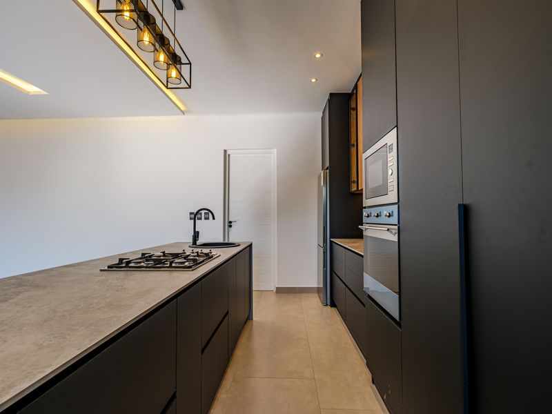 Modern kitchen featuring a sleek island with a gas hob, integrated appliances, matte black cabinetry, and contemporary recessed and pendant lighting.