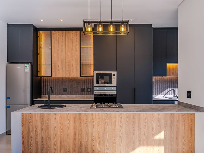 Modern kitchen featuring sleek dark cabinetry, wood-grain accents, stone countertops, integrated appliances, and stylish pendant lighting over the central island.