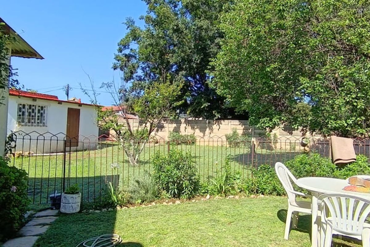A spacious rear garden featuring a lawn, mature trees, perimeter walling, and a paved area adjacent to the house.