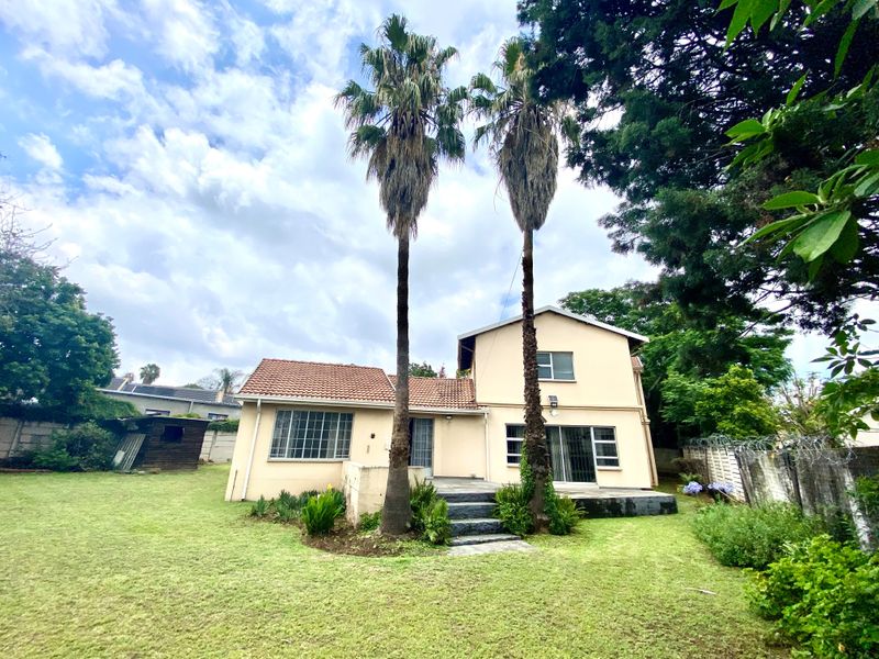 Rear view of a two-story house featuring a tiled roof, sliding glass doors, a concrete patio, and a spacious garden with mature palm trees.
