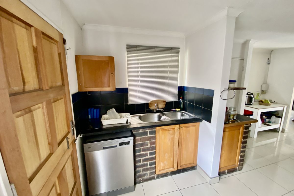 A functional kitchen area featuring timber cabinetry, a double bowl stainless steel sink, tiled splashbacks, and tiled flooring with a brick-accented base.