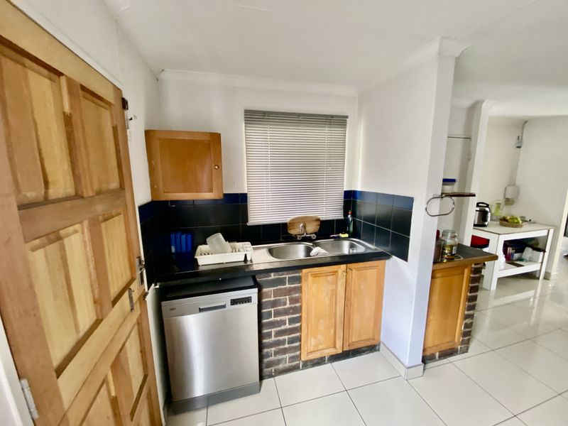 A functional kitchen area featuring timber cabinetry, a double bowl stainless steel sink, tiled splashbacks, and tiled flooring with a brick-accented base.