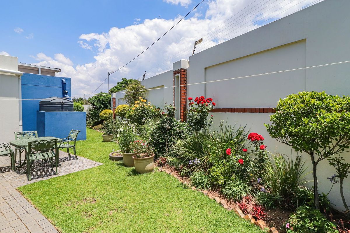 A well-maintained rear garden featuring a paved patio area, lush lawn, established flower beds, and secure perimeter walling with electric fencing.