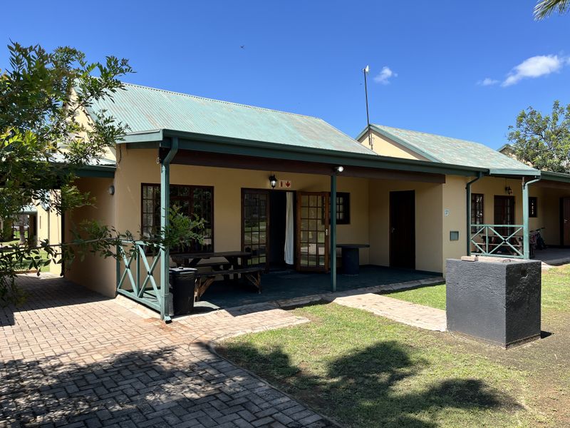 Rear view of the flat featuring a covered patio, brick-paved area, and a built-in braai, set against a well-maintained garden.