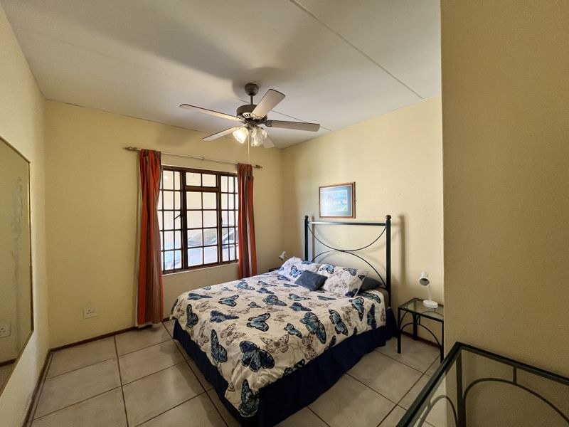 A bedroom featuring tiled flooring, a ceiling fan, and a window with security bars, providing a functional and well-lit sleeping space.
