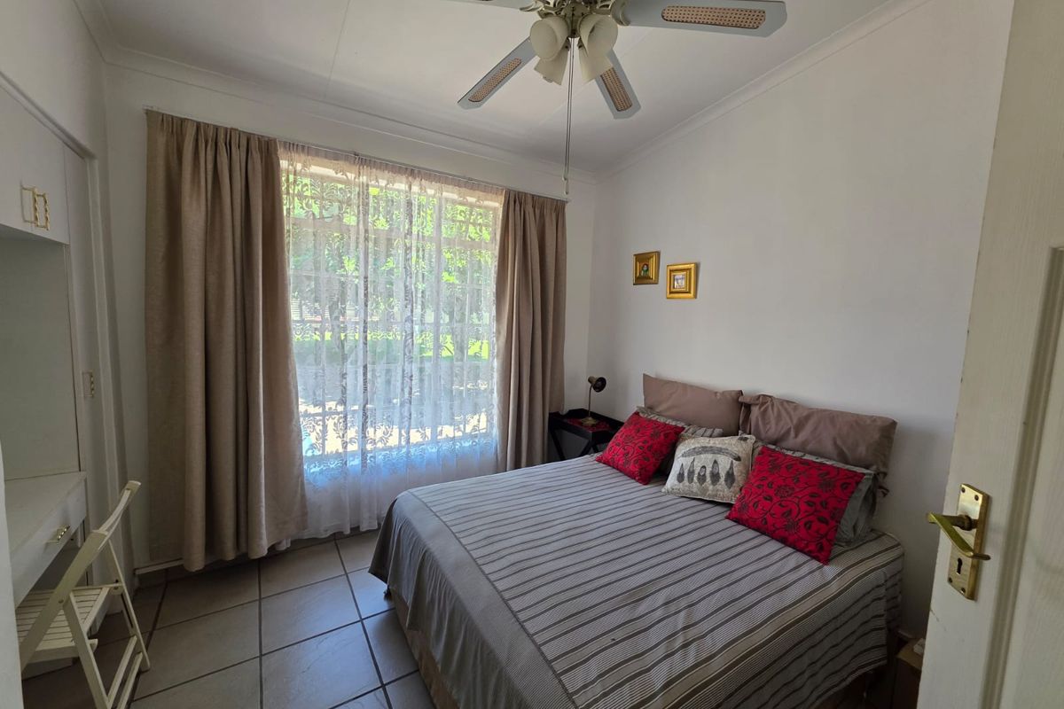 A functional bedroom featuring tiled flooring, a ceiling fan, and a large window with curtains overlooking the garden.