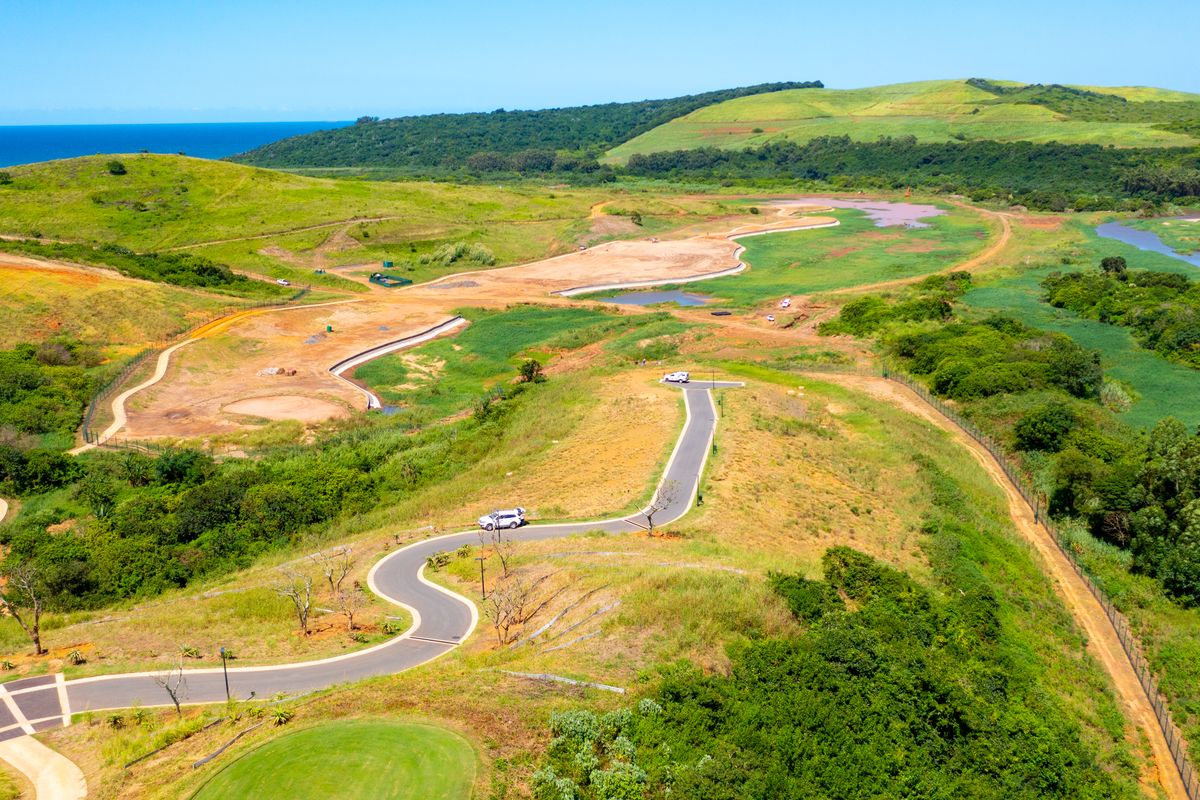 Aerial view of a residential development site featuring paved access roads, rolling green hills, and natural landscape with distant ocean views.