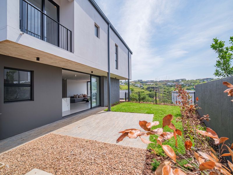 Modern townhouse rear exterior featuring a paved patio, landscaped lawn, and sliding glass doors leading to an open-plan living area with balcony above.