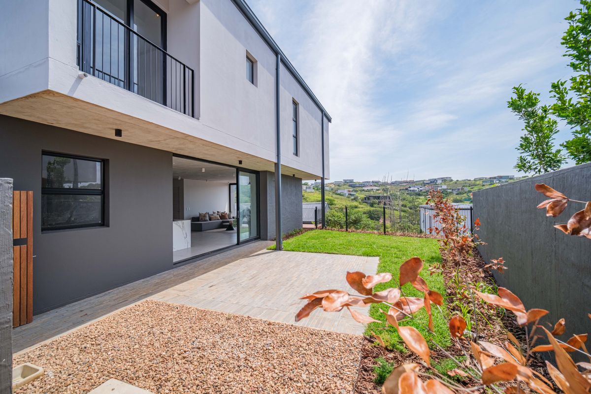 Modern townhouse rear exterior featuring a paved patio, landscaped lawn, and sliding glass doors leading to an open-plan living area with balcony above.