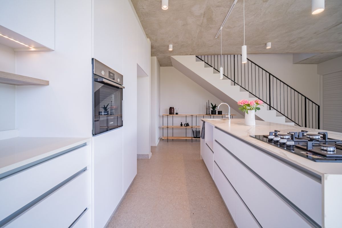 Modern kitchen featuring a central island with gas hob, built-in oven, stone countertops, and an industrial-style exposed concrete ceiling with an adjacent staircase.
