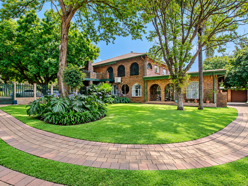 A well-maintained double-story brick house featuring arched windows, a spacious manicured lawn, and a curved paved driveway leading to the entrance.
