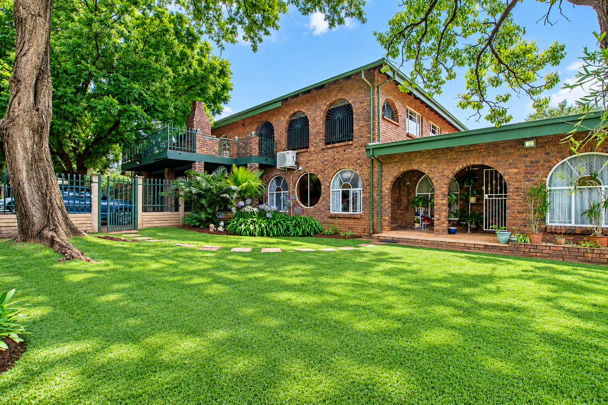A well-maintained double-story brick home featuring arched windows, a spacious upper balcony, a covered patio, and a lush, manicured garden.