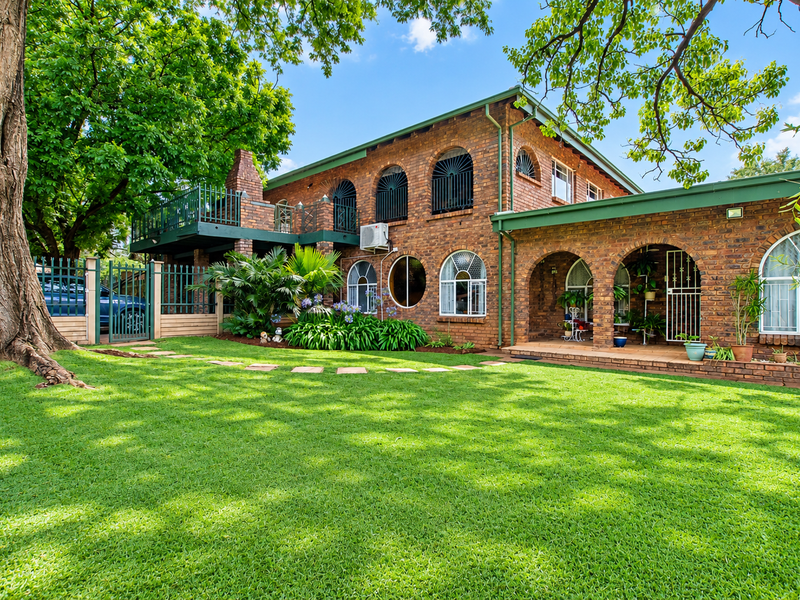 A well-maintained double-story brick home featuring arched windows, a spacious upper balcony, a covered patio, and a lush, manicured garden.
