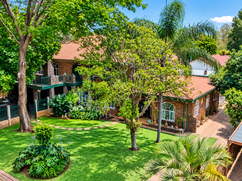 A well-maintained double-story brick home featuring a spacious landscaped garden, paved pathways, a balcony, and a covered carport area.