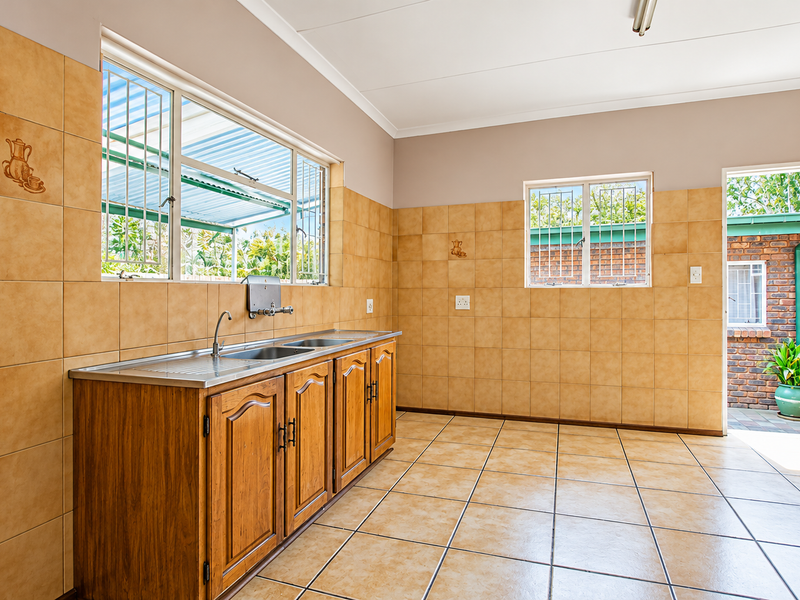 A functional scullery or utility area featuring built-in wooden cabinetry, a stainless steel sink, and ceramic tiling throughout, with direct access to the rear exterior.