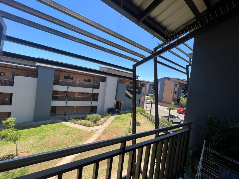 A covered balcony with a metal pergola overlooking a well-maintained apartment complex courtyard with landscaped gardens and paved pathways.