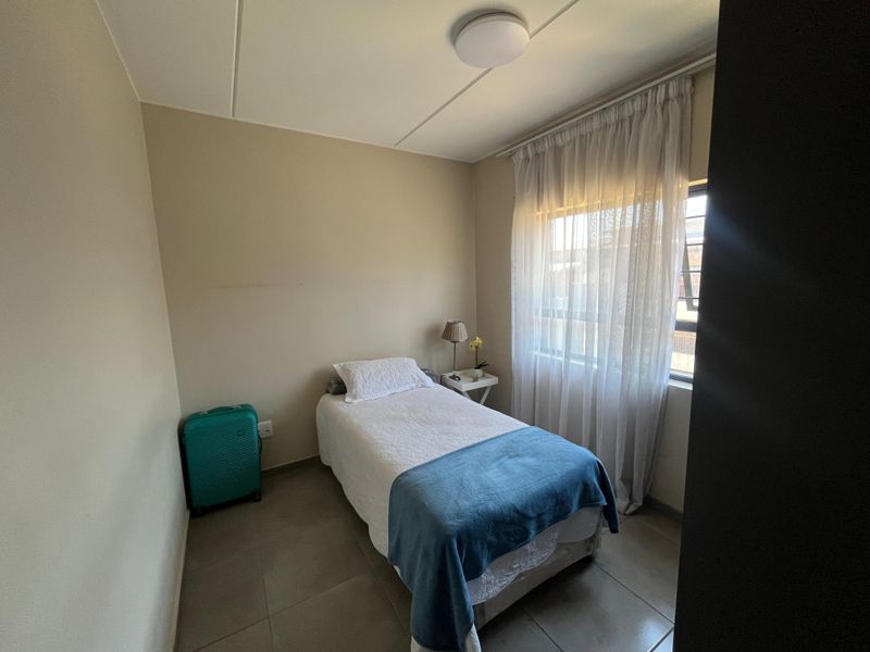 A compact bedroom featuring neutral wall tones, tiled flooring, and a window with sheer curtains, providing a functional and well-lit living space.