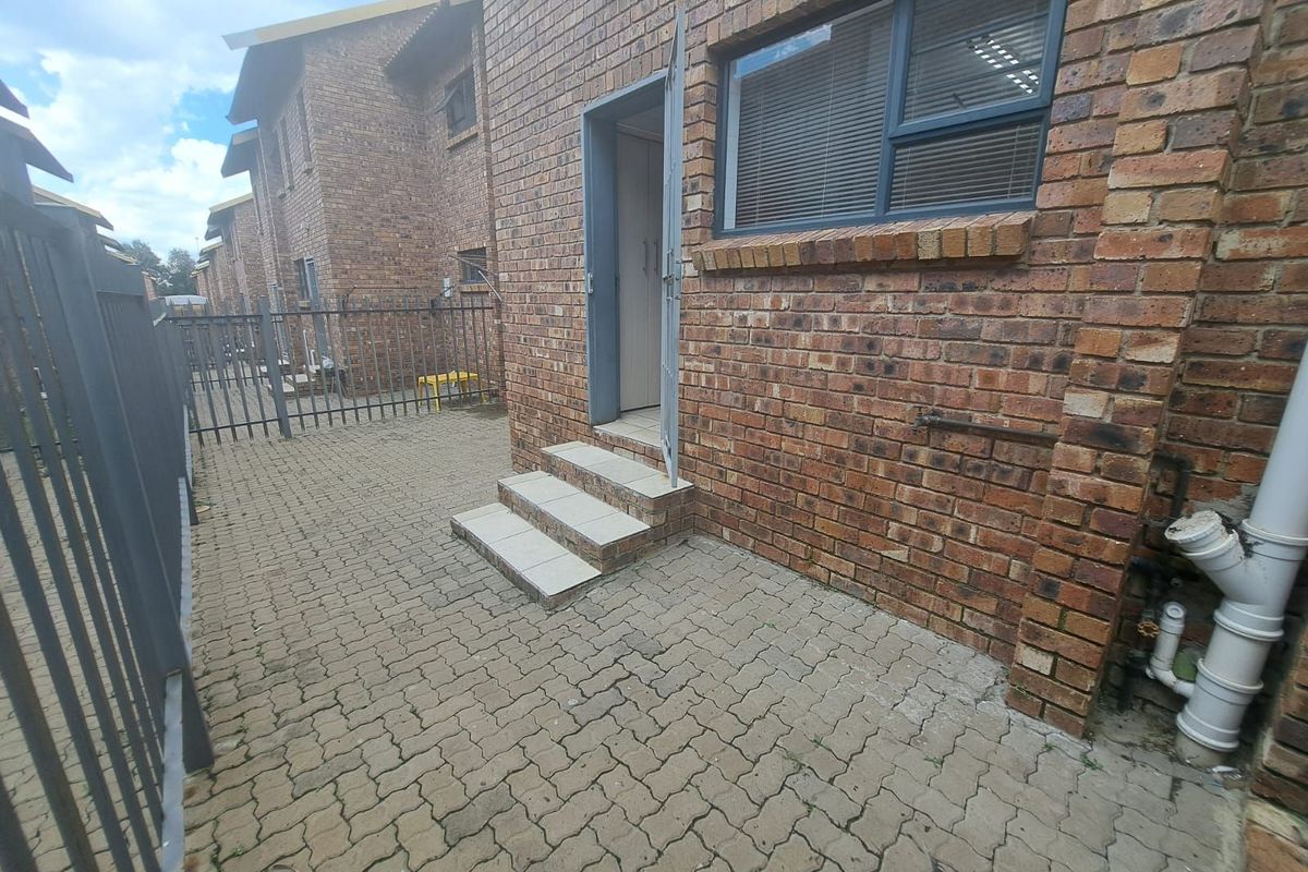Rear exterior view of a face brick apartment unit featuring a paved courtyard, security fencing, and tiled entrance steps.