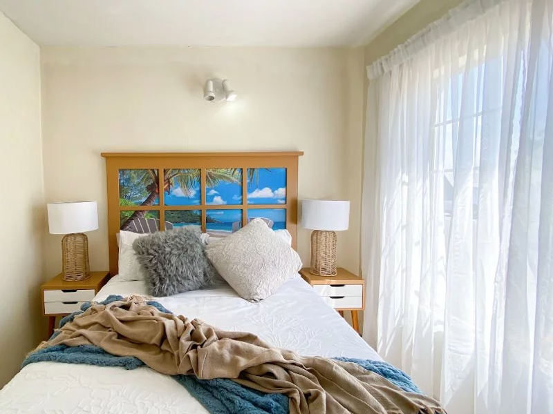 A bright bedroom featuring a wall-mounted light fixture and large window with sheer curtains, offering a clean and functional living space.