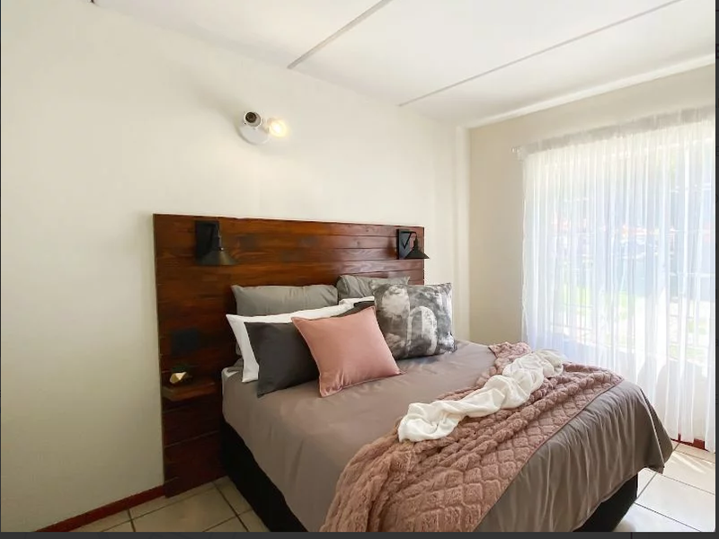 A well-lit bedroom featuring a custom built-in wooden headboard with integrated lighting and neutral tiled flooring.