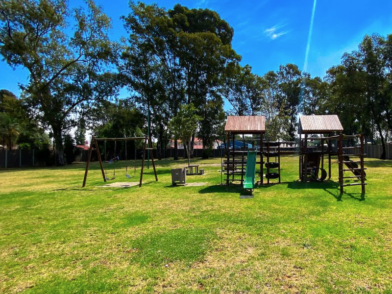 A communal outdoor playground area featuring swings, a slide, and climbing structures set within a spacious, grassed garden environment.