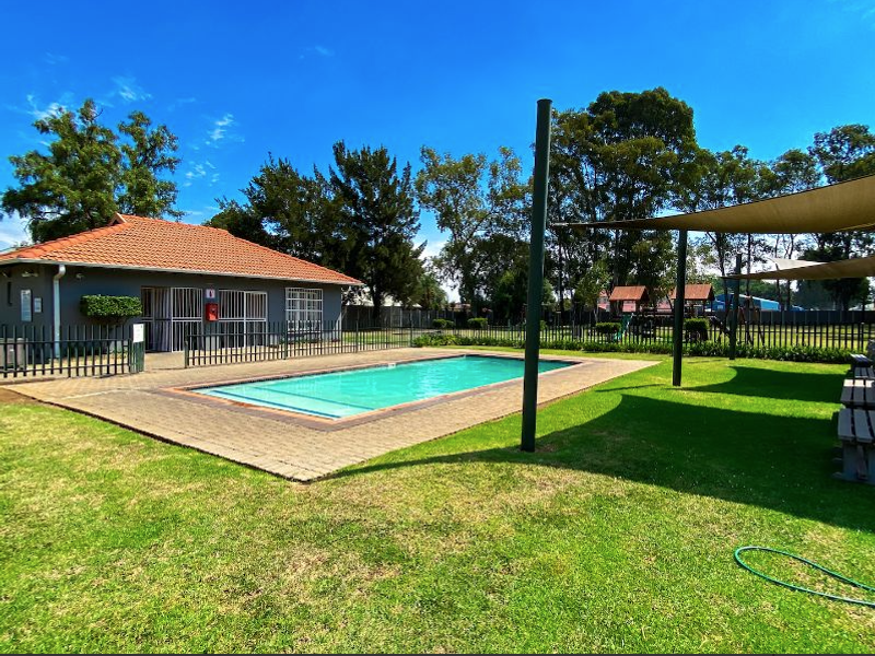 Communal outdoor area featuring a swimming pool, paved deck, and shade sail structure, set within a well-maintained garden environment.