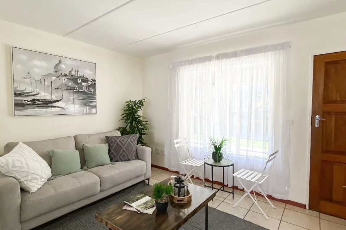 A bright living room featuring neutral tiled flooring, a solid timber door, and large windows dressed with sheer curtains.