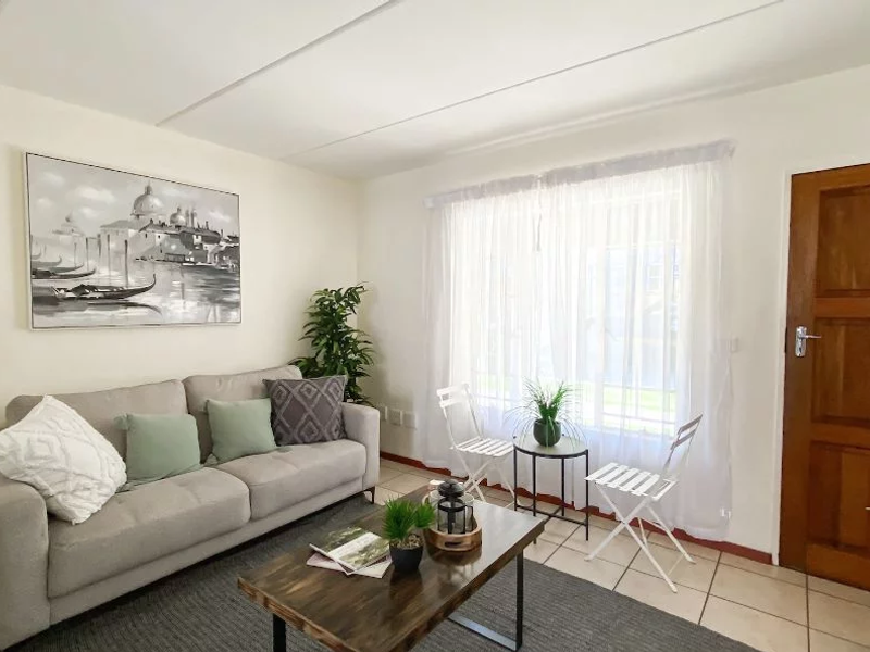 A bright living room featuring neutral tiled flooring, a solid timber door, and large windows dressed with sheer curtains.
